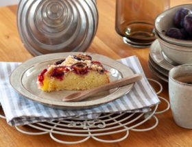 Kawałek ciasta ze śliwkami na talerzyku, kubek z kawą i miseczka ze śliwkami na wyspie kuchennej.