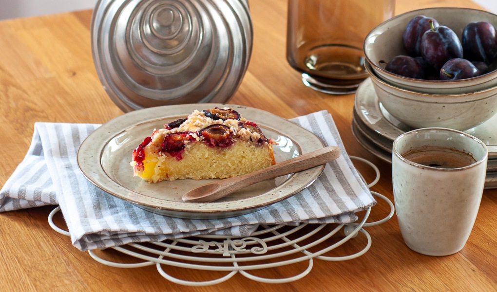 Kawałek ciasta ze śliwkami na talerzyku, kubek z kawą i miseczka ze śliwkami na wyspie kuchennej.