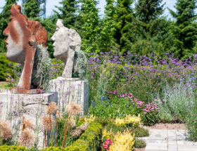Rzeźby ludzkich twarzy na postumentach wśród bujnie kwitnących kolorowych rabat w ogrodach Hortulus.