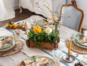 Wielkanocny stół z plecionymi podkładkami, talerzami w zielone kwiaty, lnianym bieżnikiem i drewnianą skrzynką z jaskrami.