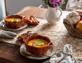 Miseczki w kształcie dyni z zupą dyniową, koszyk z kolorowymi dyniami i wazon z astrami stoją na stole.