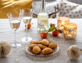 Na stole stoi talerz francuskich magdalenek, kieliszki, butelka prosecco i lampiony.