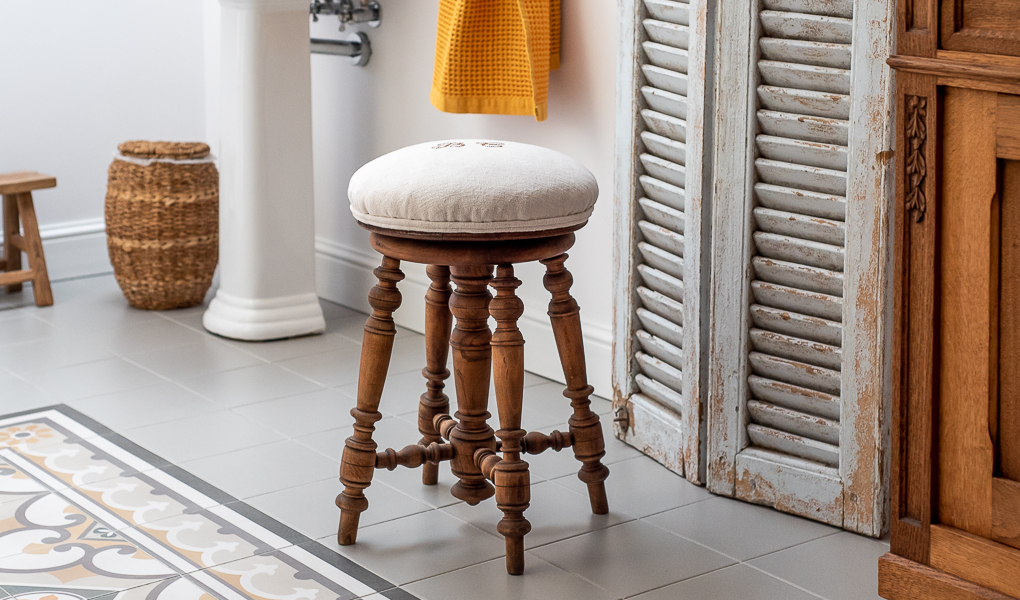Stary stołek do pianina obity kremowym lnem stoi w łazience w stylu retro.